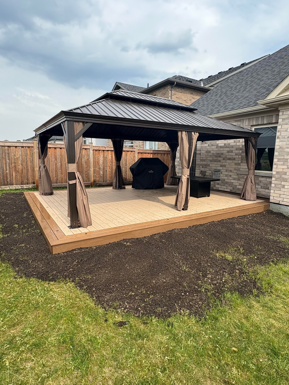 Modern backyard pergola with metal roof and tan curtains over composite deck next to brick house
