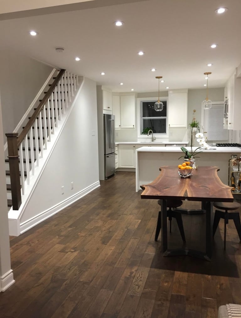 Modern open-concept kitchen and dining area with white cabinetry, wood table, dark hardwood floors, and staircase