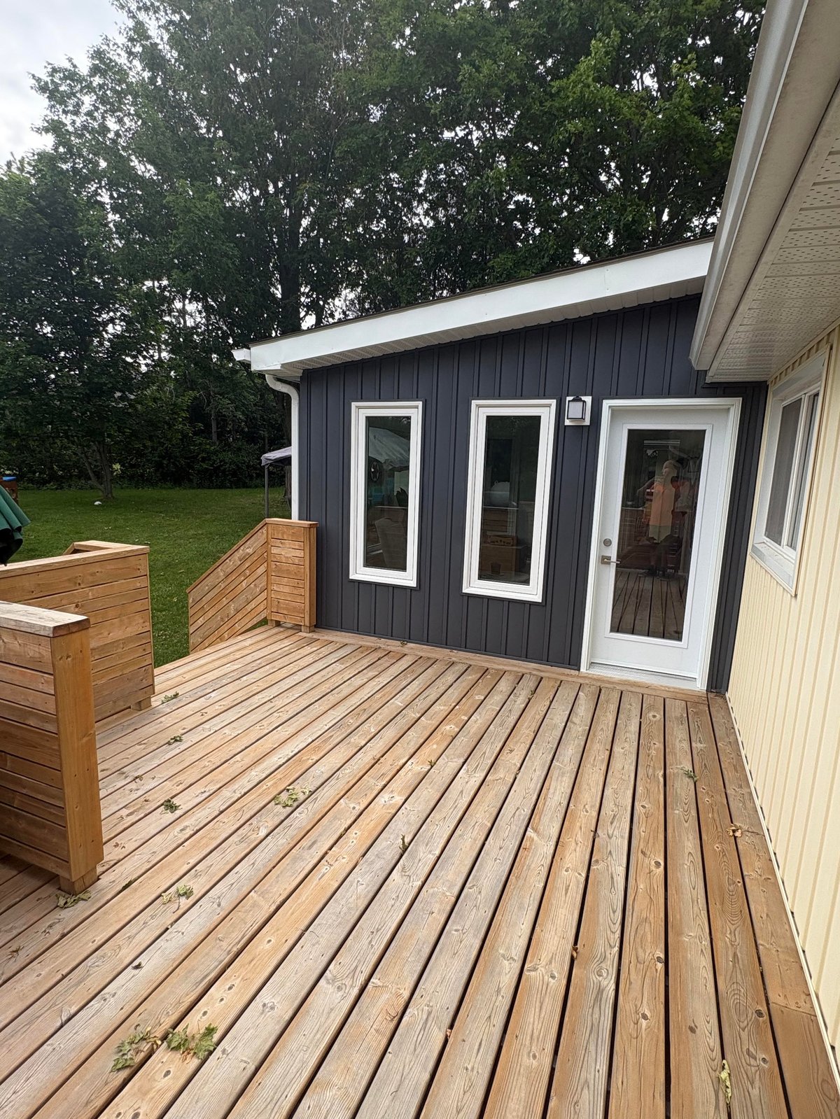 Modern dark blue and white backyard shed with white trim doors on wooden deck, surrounded by green lawn and trees