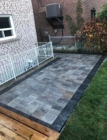 Newly installed gray paver patio in backyard with white metal railing, brick building, and lawn border