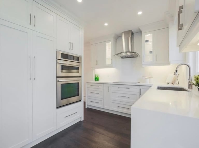 Modern white kitchen with stainless steel appliances, white cabinetry, quartz countertops, island sink, and dark wood flooring.