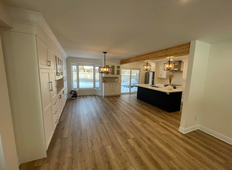 Modern open-concept kitchen and living room with hardwood floors, white cabinetry, wooden beams, and glass doors overlooking water