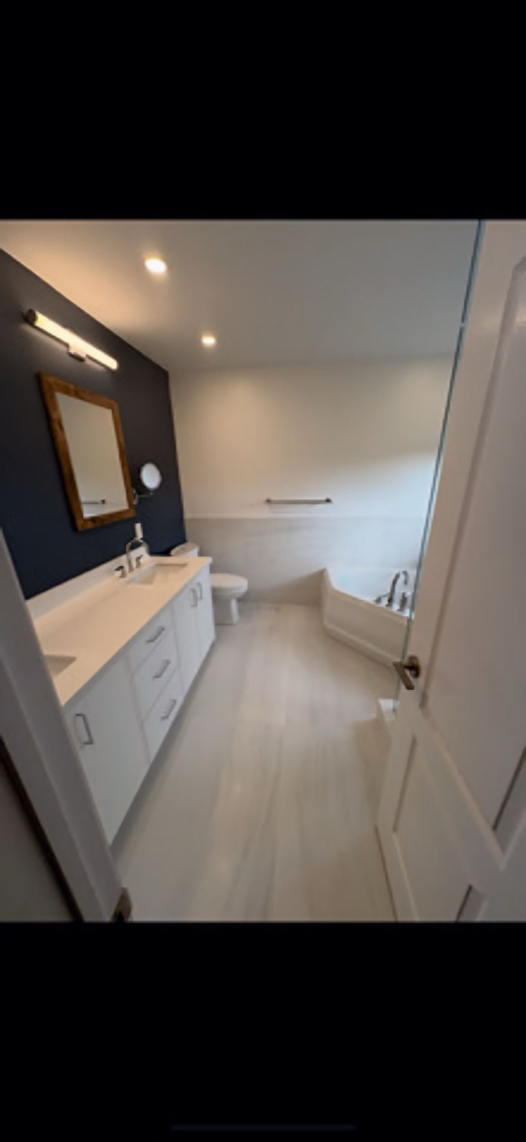 Modern bathroom interior with white vanity, dark accent wall, wooden mirror frame, and white tile flooring with bathtub