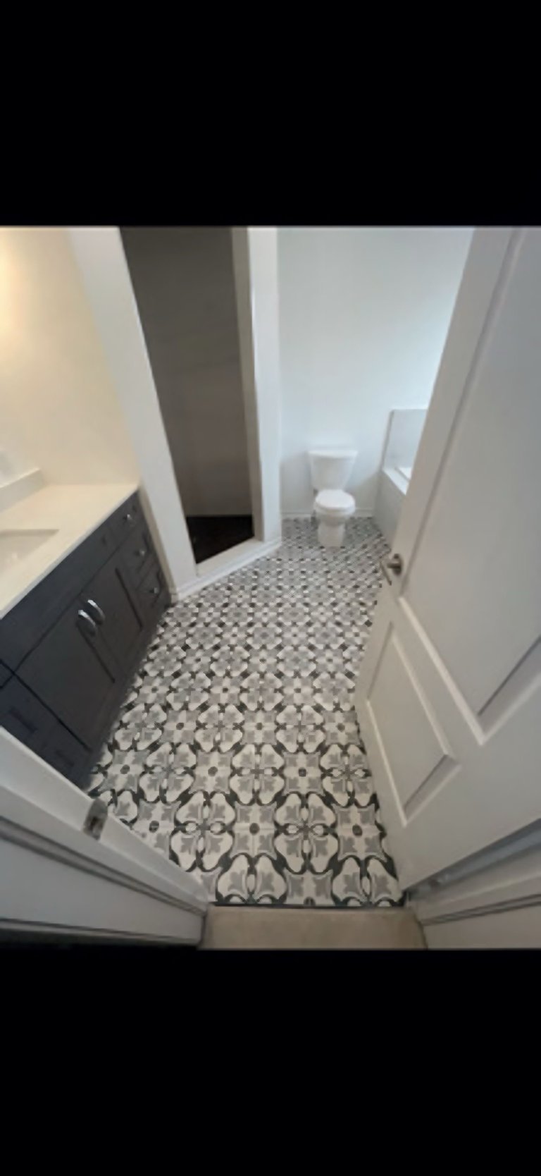 Modern bathroom with patterned tile floor, white walls, dark vanity on left, and toilet in background
