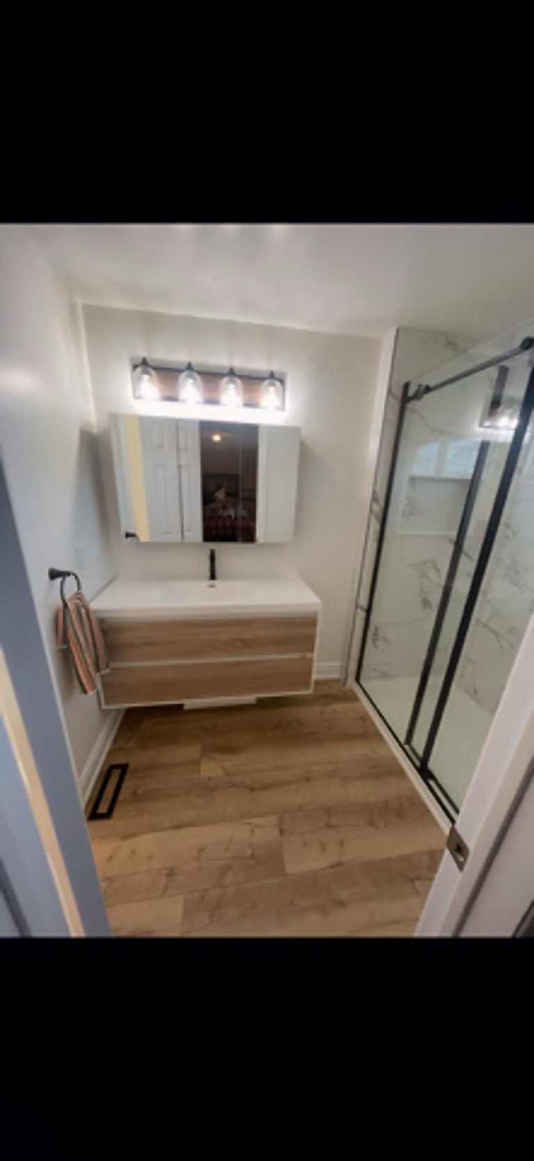 Modern bathroom with wooden vanity, mirror, wall sconces, wood flooring, and glass shower enclosure with black frame