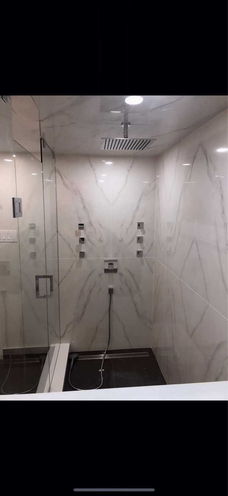 Modern walk-in shower with white marble-patterned tile walls, glass enclosure, rainfall showerhead, and multiple body jets