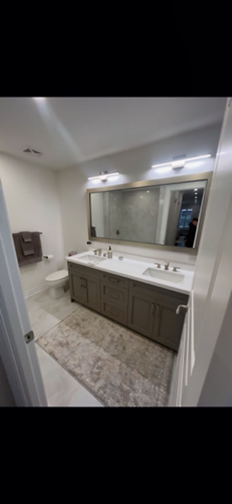 Modern bathroom with dark wood vanity, white countertop, large mirror, dual sinks, toilet, and light gray tile flooring
