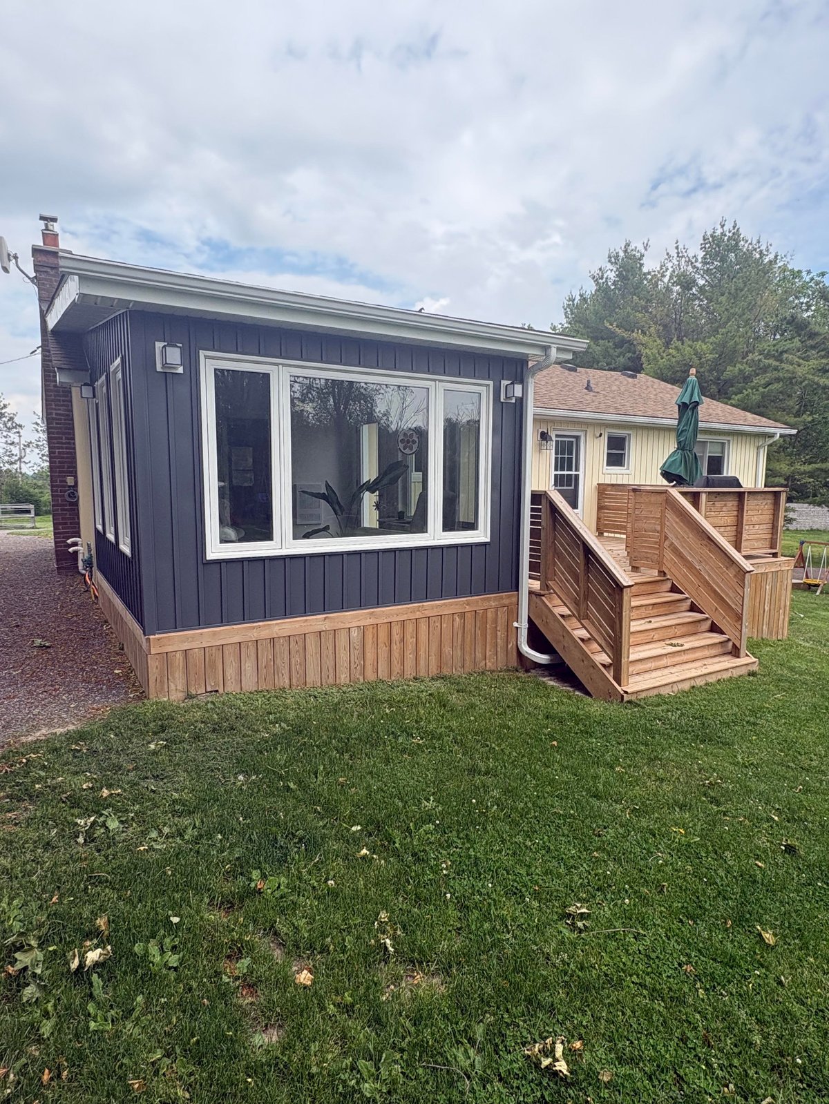 A dark blue and tan tiny house with large windows and wooden ramp entrance on a grassy lawn with buildings in the background