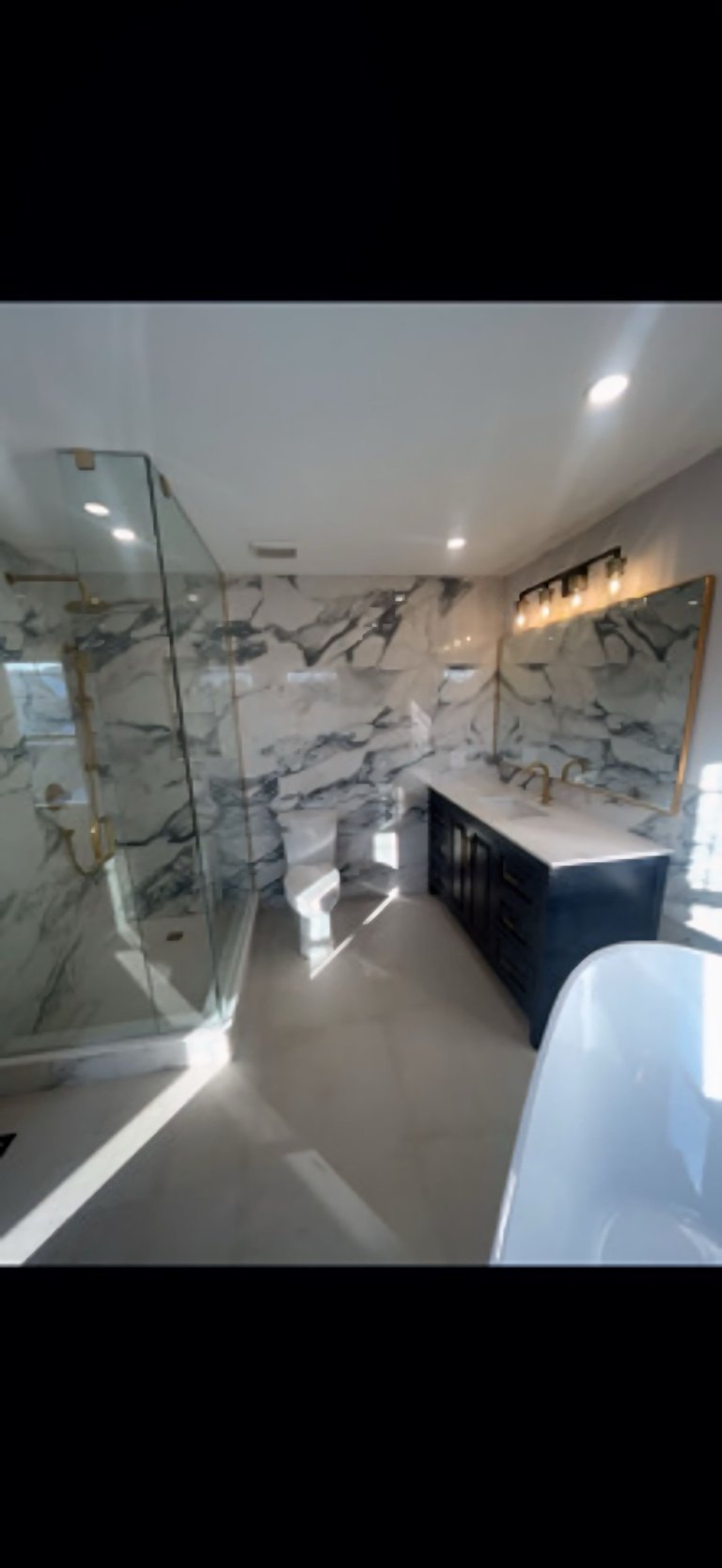 Modern bathroom with marble-patterned tile walls, a glass shower enclosure on the left, dark vanity with white countertop, soaking tub, and recessed ceiling lighting