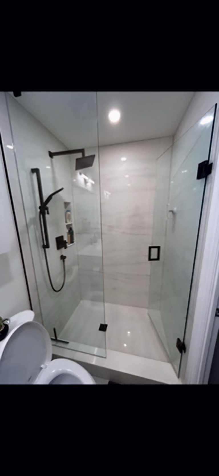 Modern bathroom with frameless glass shower enclosure, black fixtures, white subway tile walls, recessed ceiling lights, and toilet visible on left side